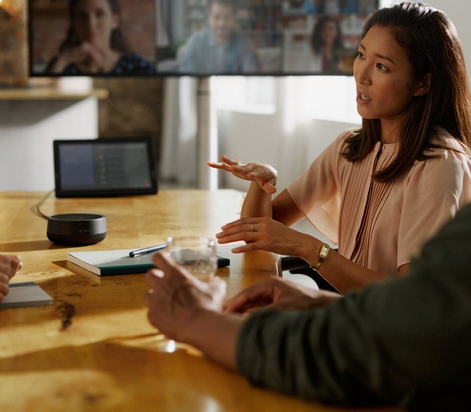 Zwei Personen im Gespräch an einem Konferenztisch mit einem Sennheiser TeamConnect Mikrofon in der Mitte, während im Hintergrund auf einem Laptop eine Echtzeit-Transkription des Meetings angezeigt wird – ideal für barrierefreie Kommunikation und Dokumentation in professionellen Arbeitsumgebungen.