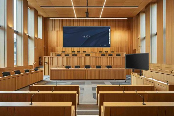 Ein leerer, moderner Gerichtssaal mit Holzmobiliar, Richterbank und großem Bildschirm für Videokonferenzen – geeignet für Videoverhandlungen nach § 128a ZPO und digitale Gerichtsverfahren.