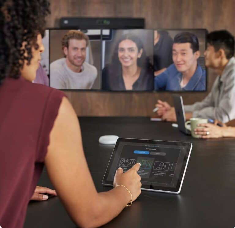 Frau steuert eine Videokonferenz mit dem Cisco Room Navigator Touch-Controller, während mehrere Personen remote auf dem Bildschirm zugeschaltet sind.