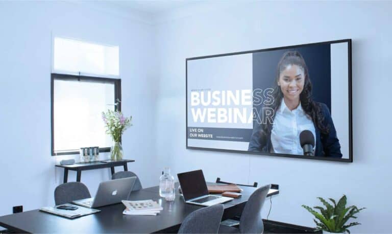 Ein Speechi Display zeigt in einem modernen Konferenzraum eine Business-Webinar-Präsentation mit einer Sprecherin im Livestream; auf dem Tisch liegen Laptops, Unterlagen und Wasser – ideal für professionelle Meetings, hybride Zusammenarbeit und visuelle Kommunikation im Unternehmen.