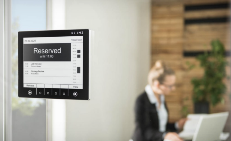 Ein ROOMZ-Display an einer Glaswand zeigt den Status „Reserved“ mit Uhrzeit und Rauminformationen an, um die Belegung von Besprechungsräumen transparent darzustellen.