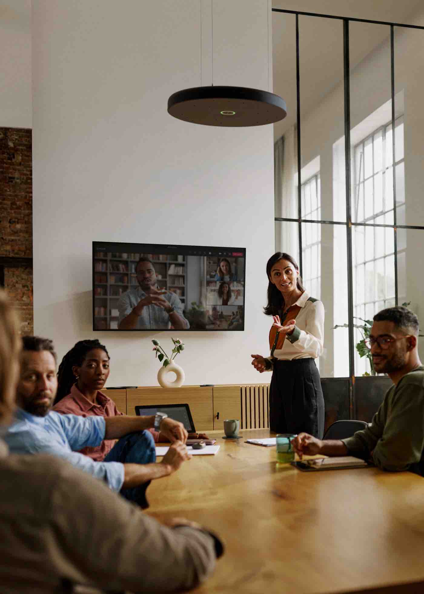 Sennheiser TeamConnect Ceiling Mic – klare Sprachqualität in mittelgroßen Meetingräumen Ein mittelgroßer Konferenzraum mit fünf Personen in einem hybriden Meeting, ausgestattet mit einem Sennheiser TeamConnect Ceiling Mikrofon, das über dem Tisch installiert ist und für hervorragende Sprachverständlichkeit trotz hoher Decken und großer Fensterfront sorgt.