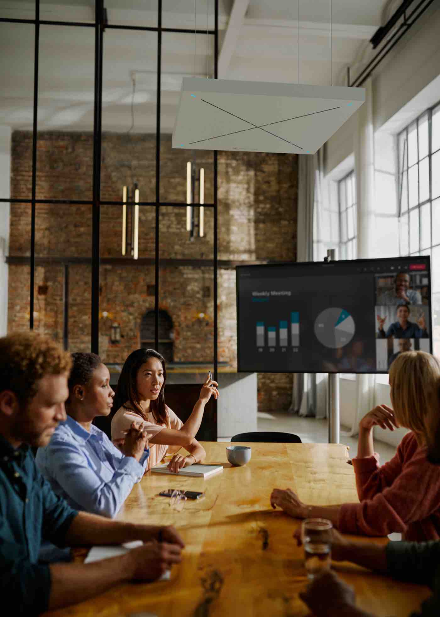 Sennheiser TeamConnect Ceiling – Deckenmikrofon im Besprechungsraum mit Fokus auf Raumakustik Moderne Besprechungssituation mit mehreren Personen an einem Konferenztisch, darüber ein installiertes Sennheiser TeamConnect Ceiling Deckenmikrofon – verdeutlicht die diskrete Integration und akustische Erfassung in professionellen Meetingräumen mit Glas- und Ziegelwänden.
