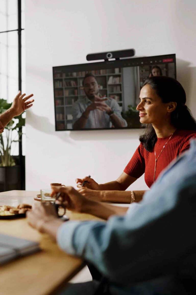 Zwei Personen sitzen an einem Konferenztisch und führen ein virtuelles Meeting, während eine Sennheiser TeamConnect Videobar S unterhalb eines großen Displays für klare Ton- und Bildübertragung sorgt – ideal für kleine Besprechungsräume und effiziente hybride Zusammenarbeit.