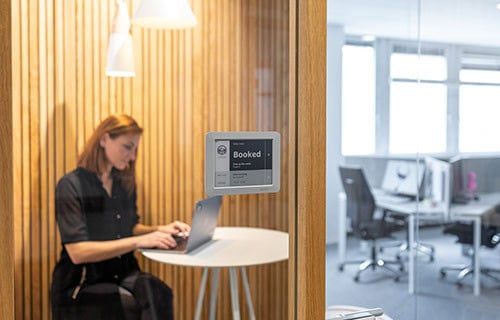 Ein Joan 6 Pro E-Ink-Türschild an einer Holzlamellenwand zeigt den Status „Booked“ für einen Meetingraum an, während im Raum eine Person am Laptop arbeitet.