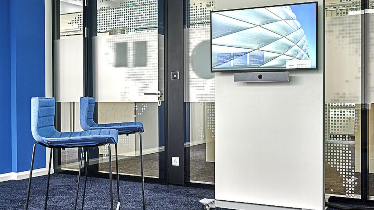 Der Fröscher Mico Slim 1 ist ein freistehender Monitorständer mit hellem Gehäuse, integriertem Bildschirm und Soundbar, platziert in einem modernen Besprechungsbereich mit Barhockern und Glastrennwänden – ideal für hybride Meetings, Präsentationen und Teamkommunikation.