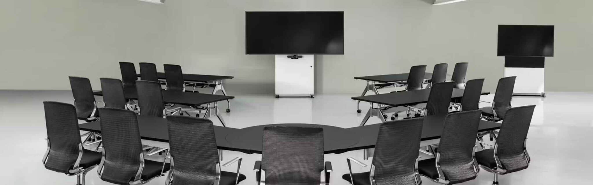 Fröscher Mico 3 Medienstele im Konferenzraum mit Monitor und Konferenztisch Die Fröscher Mico 3 Medienstele mit großem Display steht zentral an der Stirnseite eines Konferenzraums, ausgestattet mit rechteckigem Tisch und mehreren ergonomischen Konferenzstühlen – ideal für Präsentationen, hybride Meetings oder Schulungen.