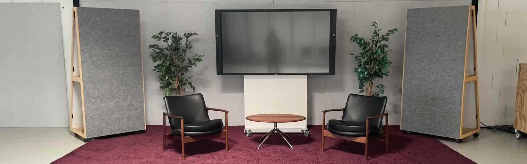 Fröscher Mico 3 Medienstele im Empfangsbereich mit Lounge-Setup Die Fröscher Mico 3 Medienstele mit großem Display steht zwischen zwei Lounge-Sesseln, umgeben von Akustikpaneelen, Pflanzen und einem Teppich – ideal für Empfangsbereiche, Wartezonen oder informelle Meetings mit digitaler Unterstützung.