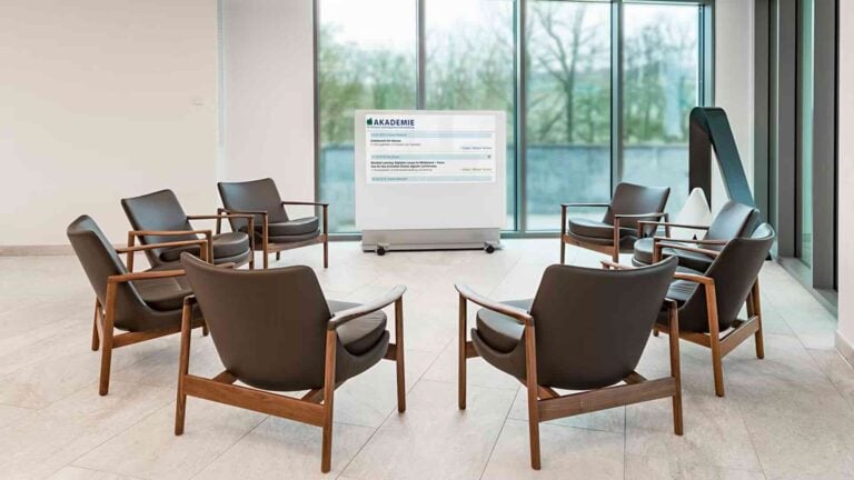 Eine mobile Medienstele Fröscher MIDS 55 mit großem Bildschirm steht in einer Lounge-artigen Sitzgruppe mit Sesseln, vor einer Glasfassade – ideal für spontane Meetings, Präsentationen oder hybride Besprechungen in offenen Bürobereichen.