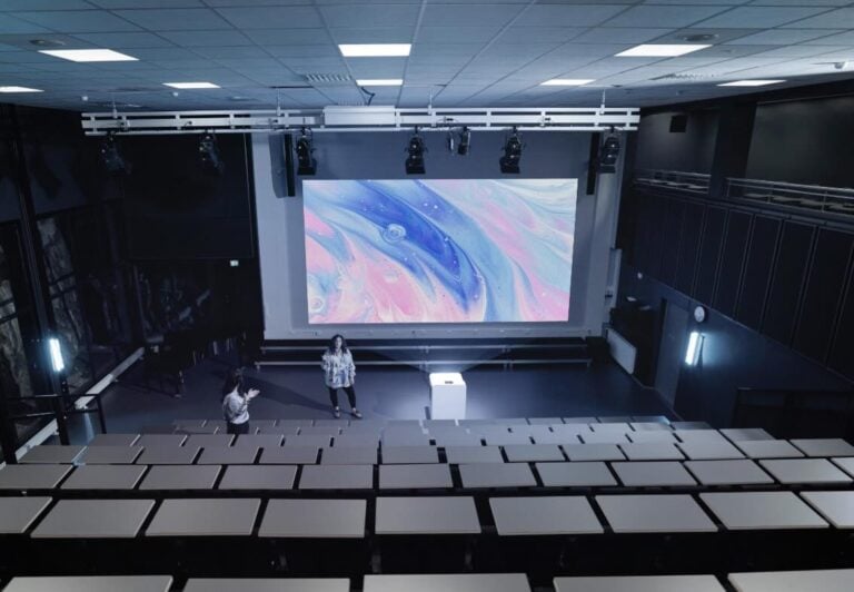 In einem großen Hörsaal ist ein Artome X20 System als mobile Präsentationslösung mit Laserprojektor, Lautsprechern und Medientechnik im Einsatz, projiziert auf eine breite Leinwand vor abgestuften Sitzreihen.