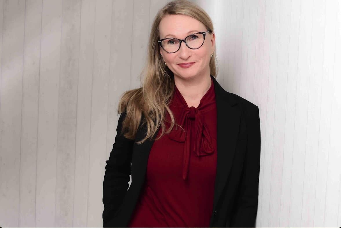 Das Bild zeigt Elgin Benkstein, Geschäftsführerin von conference-tv im Bereich Medientechnik, AV-Systemintegration und Videokonferenzlösungen, im professionellen Businessoutfit vor einer hellen Holzstrukturwand.