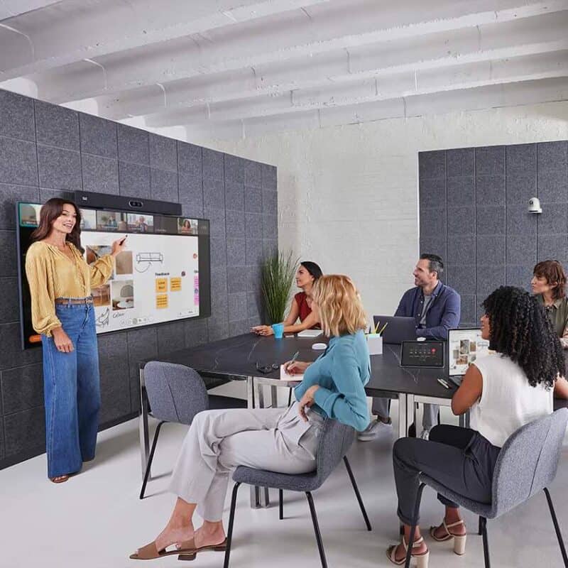 Mehrere Personen sitzen in einem modern eingerichteten Besprechungsraum vor einem großen Display mit Cisco Room Bar Pro und nehmen an einer hybriden Präsentation teil, bei der eine Person Inhalte am Bildschirm erklärt.