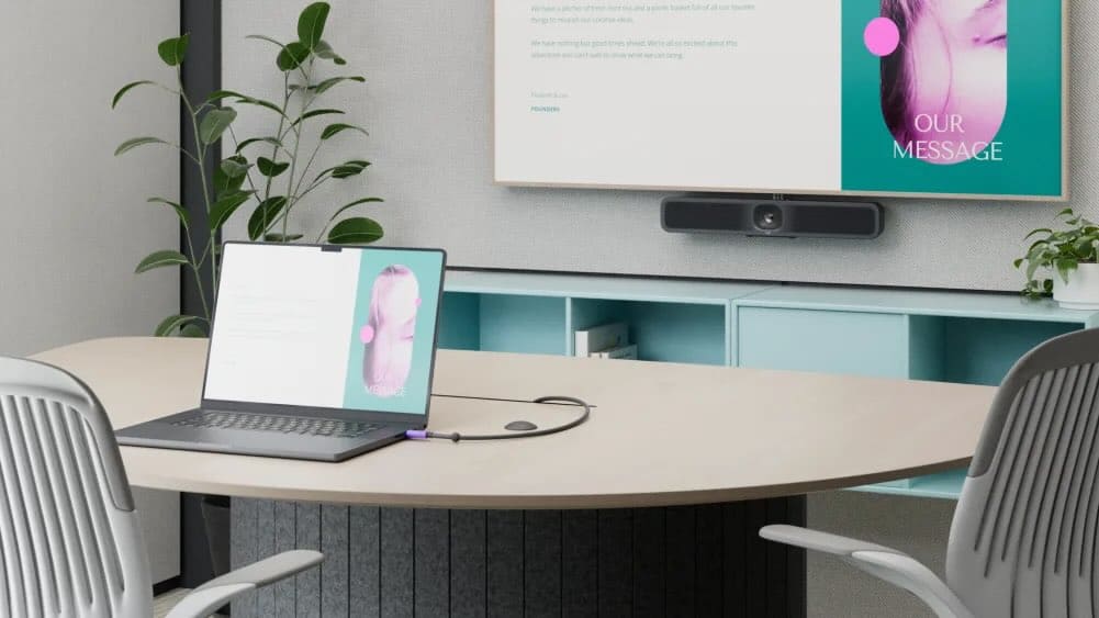 Ein moderner kleiner Besprechungsraum mit Tisch, Laptop und Logitech Videobar unterhalb eines großen Displays, auf dem Präsentationsinhalte kabellos geteilt werden – ideal für Videokonferenzen, Huddle Rooms und hybride Teamarbeit.