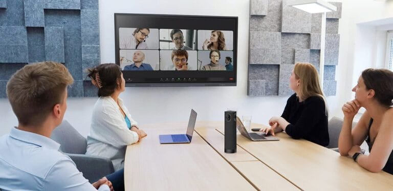 Fünf Personen sitzen in einem modernen Konferenzraum um einen Tisch, in der Mitte steht eine Speechi 360°-Konferenzkamera, während auf einem großen Bildschirm eine Videokonferenz mit mehreren zugeschalteten Teilnehmern läuft. Die Szene zeigt den realen Einsatz der Kamera in hybriden Meetings.