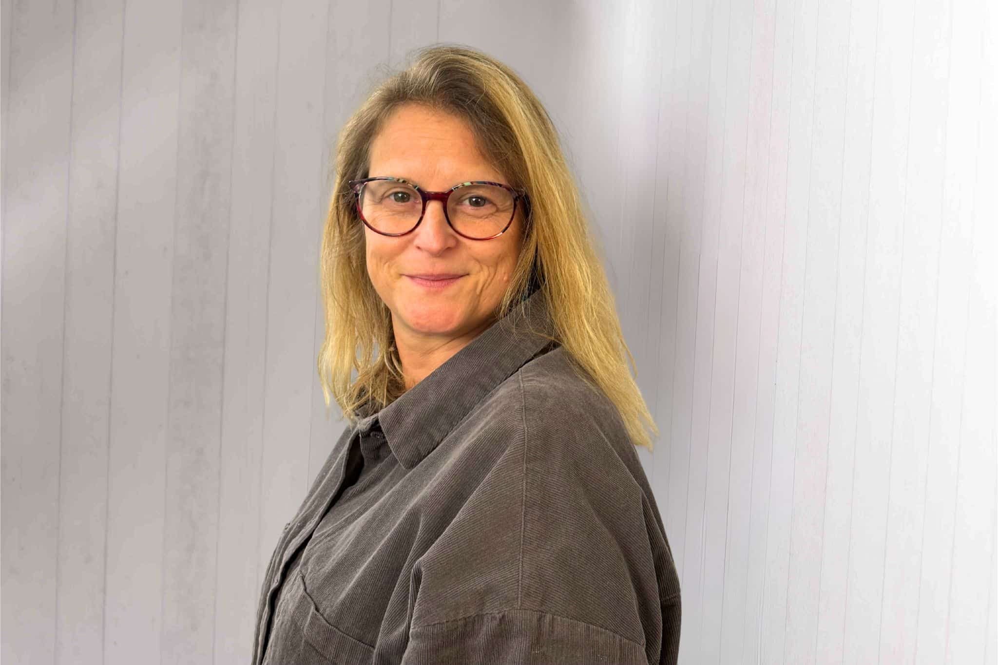 Das Porträt zeigt Claudia von Selzam, zuständig für Buchhaltung und Rechnungswesen bei conference-tv, vor einer hellen Wandstruktur in grauer Jacke und mit Brille.