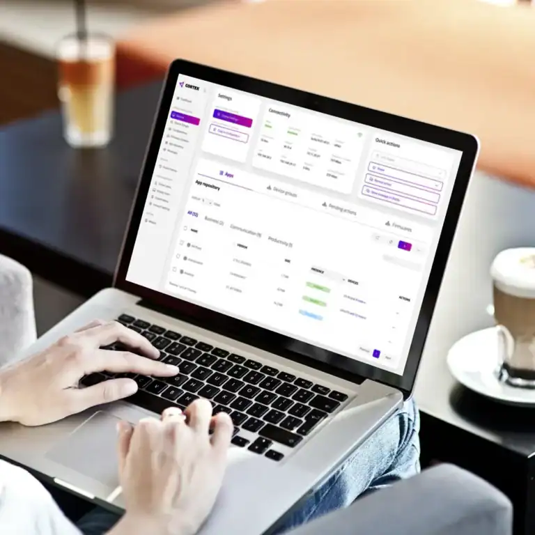 Eine Person bedient einen Laptop auf einem Tisch, auf dem Bildschirm ist eine farbige Benutzeroberfläche sichtbar. Rechts steht eine weiße Kaffeetasse.
