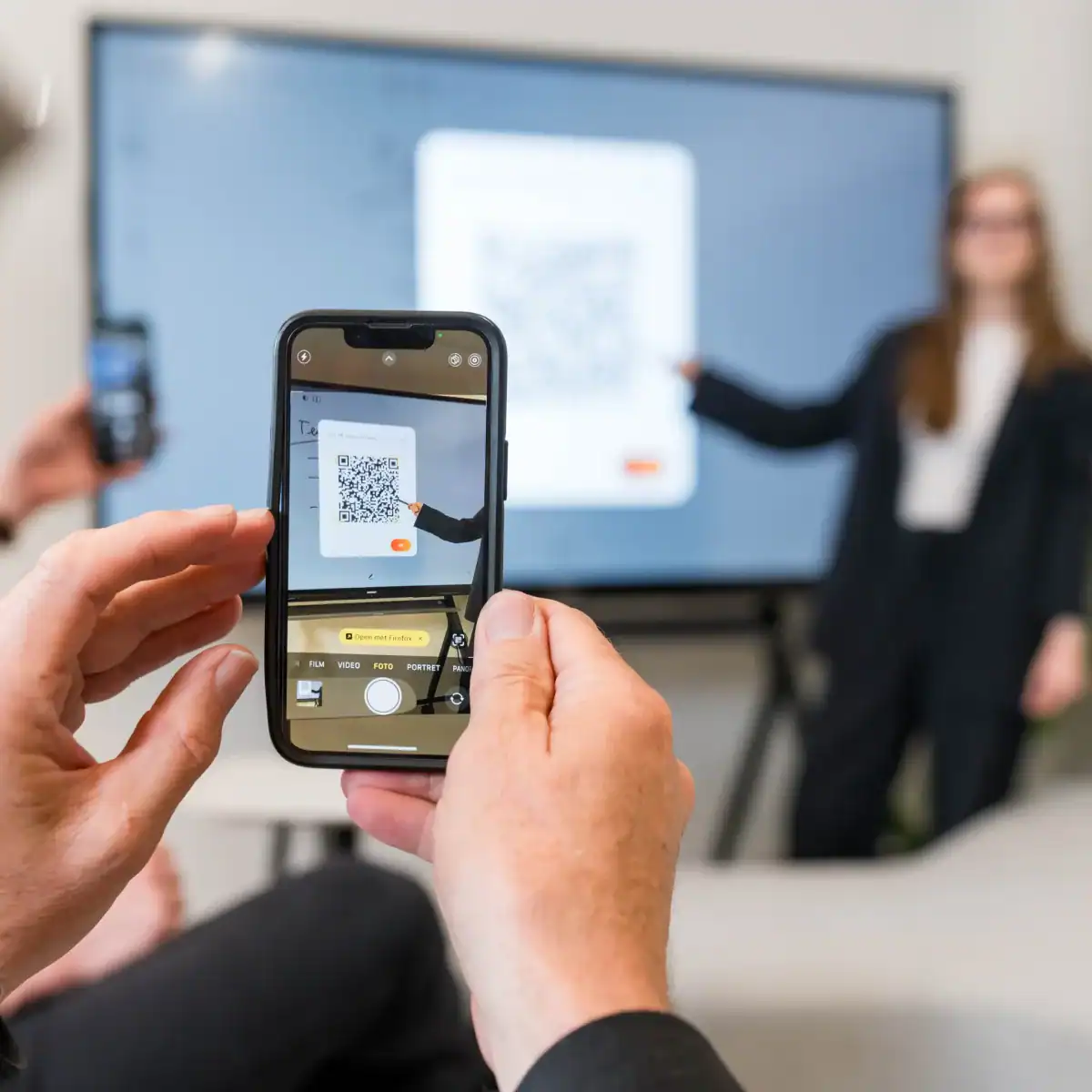 i3CONNECT Cortex QR-Code-Zugriff am Touchdisplay Eine Hand hält ein Smartphone vor einen großen Bildschirm und scannt einen QR-Code; dahinter steht ein interaktives Display mit einer Person im Hintergrund.