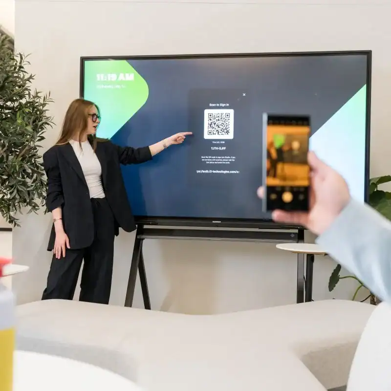 i3CONNECT Studio Präsentation am Touchdisplay Eine Person steht vor einem großen Touchdisplay und zeigt auf den Bildschirm; im Vordergrund hält eine Hand ein Smartphone vor das Display.