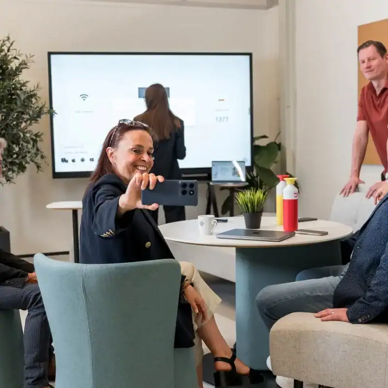 i3CONNECT Studio Besprechung am Touchdisplay Mehrere Personen sitzen und stehen in einem Besprechungsraum; im Hintergrund ist ein großes Touchdisplay sichtbar, auf einem Tisch stehen Flaschen, Becher und Geräte.