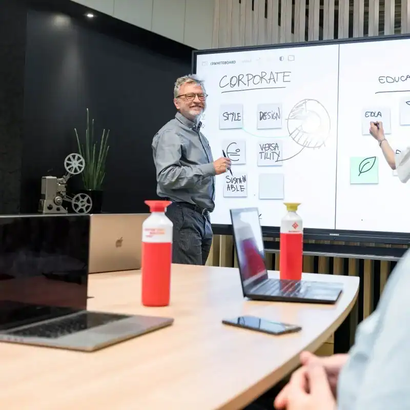 i3-connect-studio-whiteboarding Eine Person steht vor einem großen Touchdisplay im Besprechungsraum; auf dem Bildschirm sind Whiteboard-Inhalte sichtbar, im Vordergrund stehen Flaschen und ein Laptop auf dem Tisch.