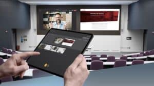 Hände halten ein Tablet vor einem Hörsaal mit zwei großen Bildschirmen, auf denen unterschiedliche Inhalte gleichzeitig angezeigt werden.