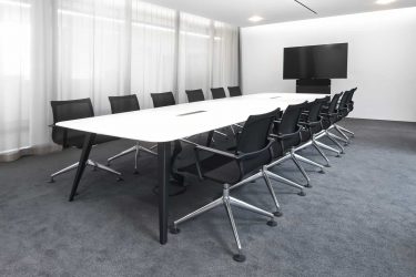 Ein heller Besprechungsraum mit einem langen Holzmedia C12 Konferenztisch, schwarzen Konferenzstühlen und wandmontiertem Display eignet sich für Meetings, Workshops und Videokonferenzen in professionellem Umfeld.
