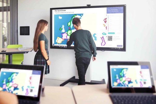 Zwei Schüler stehen vor einem großen Touchdisplay im Klassenraum; im Vordergrund stehen zwei Tablets, auf deren Bildschirmen bunte Lerninhalte sichtbar sind.