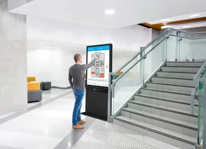 Ein Mann nutzt in einer modernen Lobby einen interaktiven Sony TEOS Digital-Signage-Bildschirm neben einer Treppe, um digitale Wegweiser und Raumpläne für die Orientierung im Gebäude abzurufen.