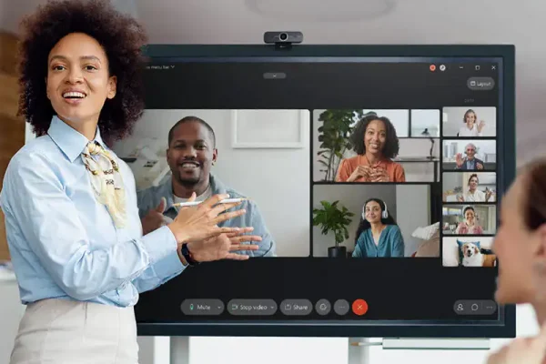 Eine Frau präsentiert vor einem großen Bildschirm mit aktiver Videokonferenz, auf dem mehrere Gesprächsteilnehmer zugeschaltet sind; die Speechi SPE-MG-104 Full-HD-Kamera ist oberhalb des Displays montiert – geeignet für kleine Meetingbereiche und hybride Zusammenarbeit.